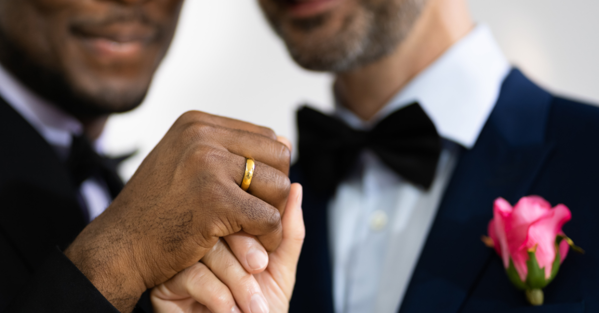 Two men holding hands in tuxedos with wedding rings on